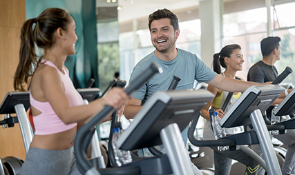 Two people using cardio machines, talking, and smiling in a gym cardio floor